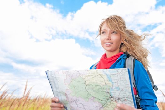 Young pretty woman tourist standing in wheat field with map. Blue cloudy sky in background behind girl. Tourism travel and hiking outdoor in summer. Healthy lifestyle.