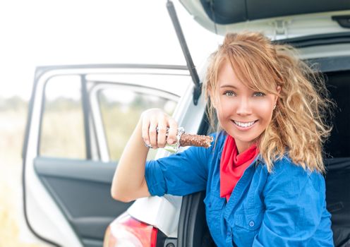 Pretty woman sitting in trunk and smiling. Portrait of young girl with beautiful hairstyle holding chocolate bar. White car with open doors and happy blonde looking into camera. Outdoor eating.