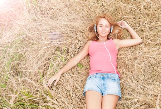 Beautiful girl relaxing in headphones outdoors. Pretty smiling woman with closed eyes listening to music lying on hay in field. Harmony of human and nature. Countryside.