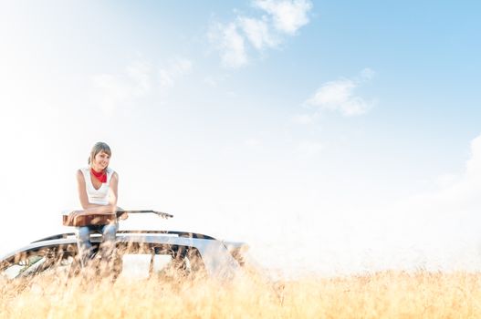 Young smiling girl sitting on her car with guitar in hands with wheat field in foreground and blue sky in background behind woman. Freedom and music. Active leisure and hobby.