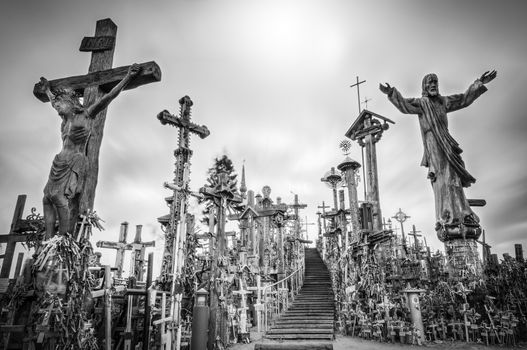 Hill of crosses in Siauliai, the city of Lithuania. Old stairway goes to gray sky. Stairs lead to the top of hill. Crosses and statues of saints near. Sun shines weakly, so place looks mysterious.