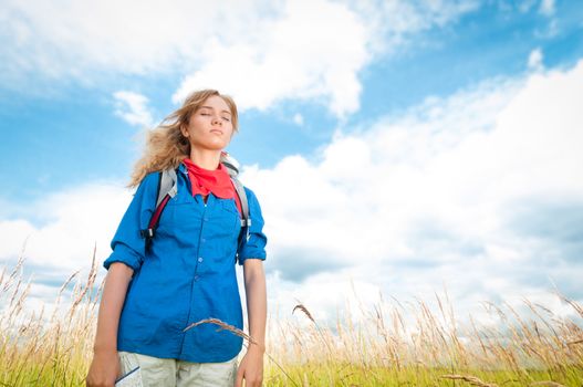 Pretty young tourist woman with closed eyes enjoying fresh air in wheat field with blue cloudy sky in background. Tourism travel and hiking outdoor in summer.