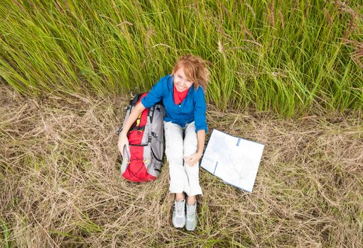 Young pretty female tourist with backpack and map having stop in field. Happy and smiling girl on halt sitting on dry grass. Tourism travelling and hiking outdoors in summer.Healthy lifestyle.