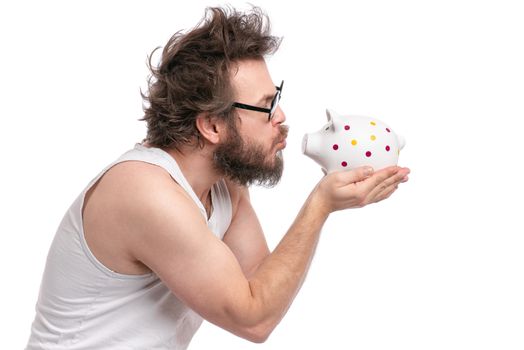 Crazy bearded Man with funny Haircut in eye Glasses holding Piggy Bank, isolated on white background. Saving Money concept. Male carefully kisses his piggybank.