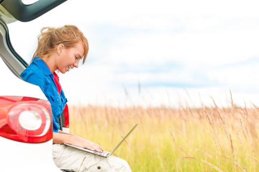 Young happy girl sitting and working on computer. Beautiful woman near car in meadow holding laptop. Person working outdoor. Tail light of car in foreground and field with sky in background.
