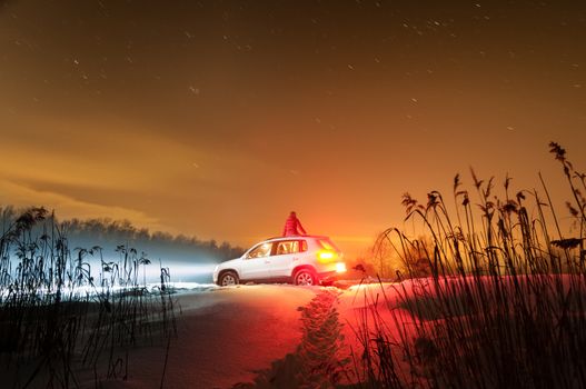Starry winter night. White car with woman sitting on it. Headlights give blue and red light. Snow and grass in foreground and beautiful sky in background.