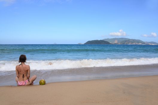 NHA TRANG, VIETNAM – 29 FEBRUARY 2020 : Sexy bikini body woman sitting on nha trang  beach behind is an Vinpearl Land