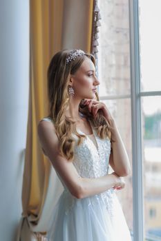 Close-up portrait of the bride in a white wedding dress by the window, looks attentively
