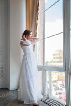 A bride in a white wedding dress straightens her hair while standing by a huge floor-length window.