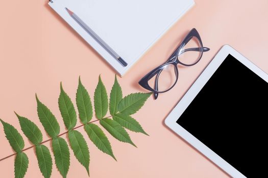 Tablet mockup on beige work desk surrounded with glasses, notepad, plant and pencil. Free space beside for text. Flat lay composition.