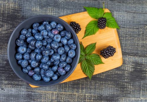 blueberry berry in dark gray ceramic bowl on wooden cutting board on dark blue wooden background. top view soft focus