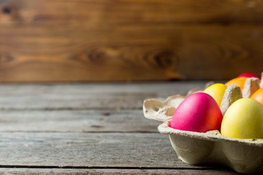 Dyed easter eggs in cardboard box on wooden background. Easter background with space for a text.