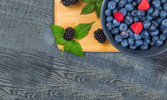 blueberry berry in dark gray ceramic bowl on wooden cutting board on dark blue wooden background. top view soft focus