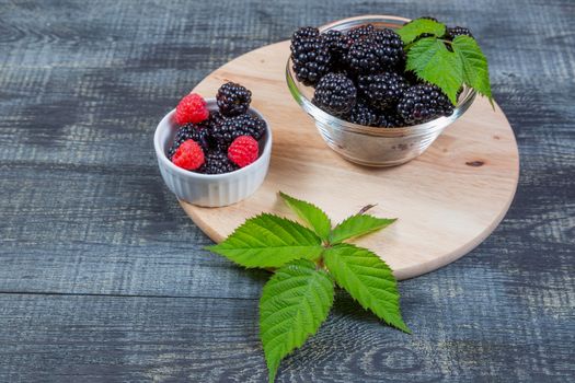 ripe blackberry with leaves on a wooden cutting board in a glass bowl on dark blue wooden background.