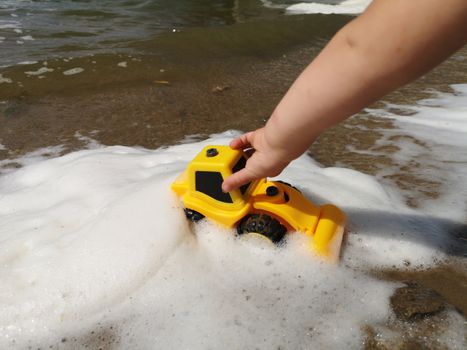  A hand of little boy with a bright yellow toy car in the foam of the lake. Summer kids holiday concept. Rest on the shore.