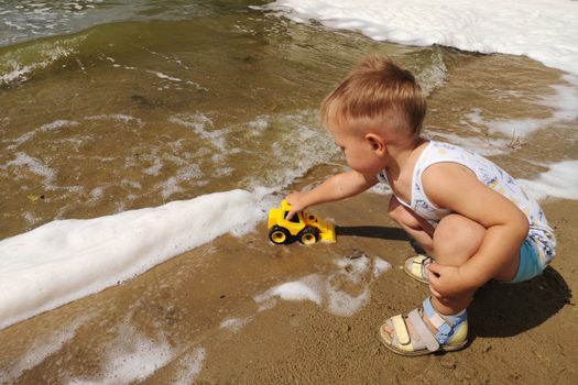 A cute little boy plays on the shore of the lake with a bright yellow toy car in the foam of the lake.Summer kids holiday concept. Rest on the shore.
