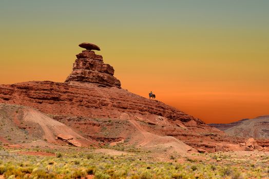 A cowboy on horseback passes by Mexican Hat Rock in Mexican Hat, Utah, USA