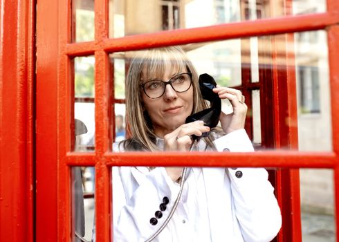 A woman wearing glasses, a white coat, jeans and a white shirt calling from a red phone box in the city center