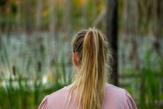 young caucasian woman standing back to camera in evening swamp with blurred dry trees and cattai in the background.