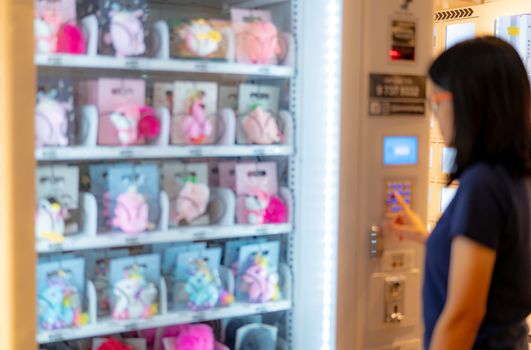 Blur Asian woman pressing button on vending machine to buy doll. Female tourists buying doll at automatic vending machine in airport shopping mall. Self-service device. Consumer and vending machine.