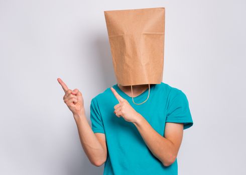 Teen boy with paper bag over head pointing finger away at copyspace, on gray background. Teenager cover head with shopping bag pointing finger at something. Child pulling paper bag over head