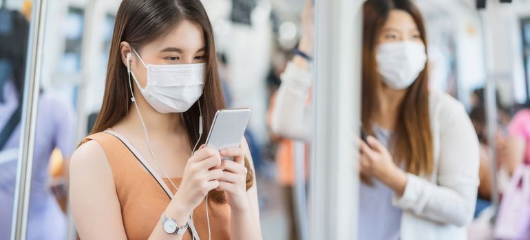 Young Asian woman passenger wearing surgical mask and listening music via smart mobile phone in subway train when traveling in big city at Covid19 outbreak, Infection and Pandemic concept
