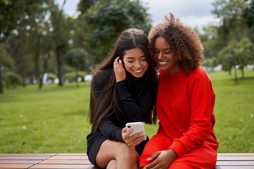 Diversity and feminity concept. Happy smiling Confident young mixed race women sitting bench park outdoor Asian and african american female resting on nature summer day using smartphone.
