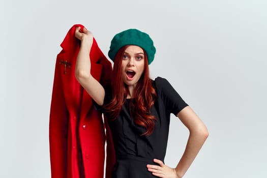 A woman holds in her hand a red coat and a dark T-shirt Green hat cropped view of a light background. High quality photo