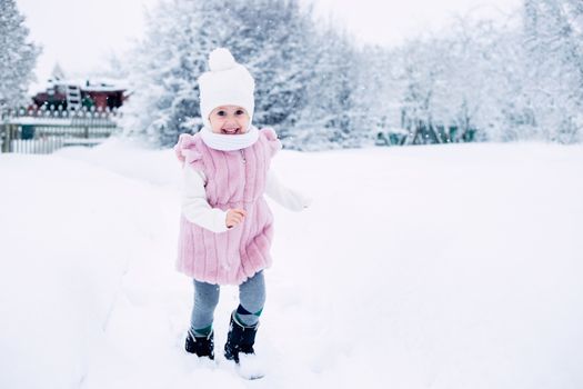 .A little girl in a pink fur coat stands in the middle of a snow-covered park on a cloudy winter day