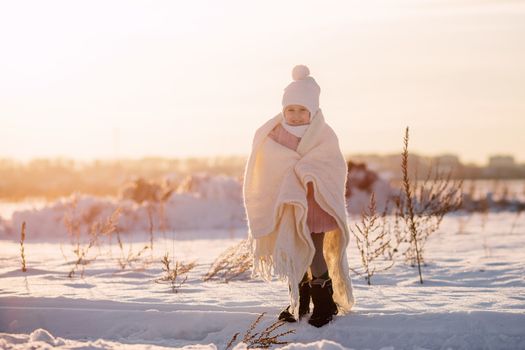 .A little girl stands wrapped in a blanket in the middle of a field in winter at sunset