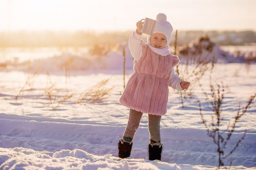 .A little girl stands with a mug of hot chocolate in the middle of a field in winter in the setting sun