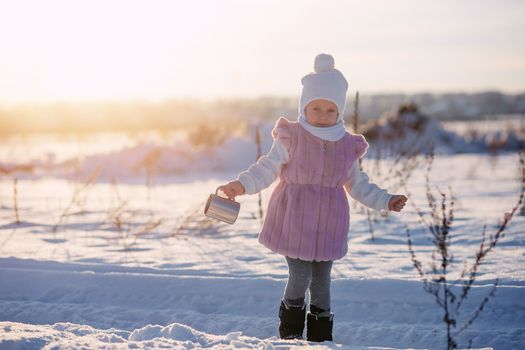 .Little girl stands with a mug in the middle of a field in winter at sunset