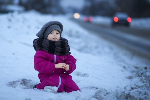 .A little girl sits in the evening on the snow by the road and watches passing cars while waiting for her dad to come home from work