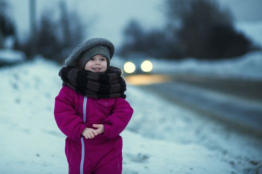 .A little girl stands in the snow by the road in the evening and watches passing cars while waiting for her dad to come home from work