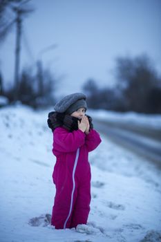 Little girl warms her hands with her breath on a winter evening near the road