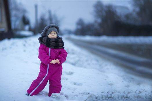 .A little girl stands in the snow by the road in the evening and watches passing cars while waiting for her dad to come home from work