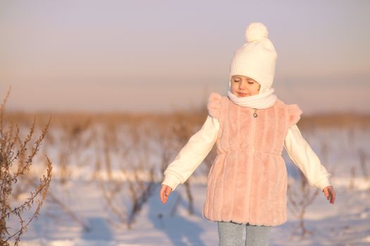 A little girl stands in the middle of a field in winter at sunset in the rays of the setting sun