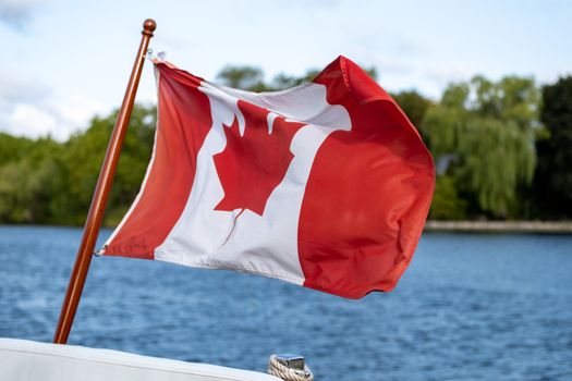 A Canadian flag flies in the wind at the stern, or back, of a small boat. Trees on a free. shore and the blue water of a river are seen behind it.