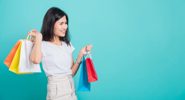 Portrait happy Asian beautiful young woman standing wear white t-shirt, She holding shopping bags multi color on hand and looking to space, shoot photo in a studio on a blue background