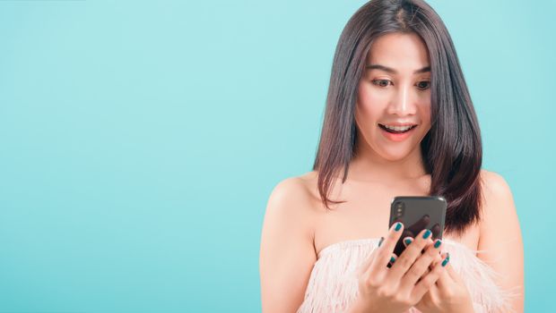 Asian happy portrait beautiful young woman standing smile her  holding smartphone or mobile phone and looking to the phone on blue background with copy space for text