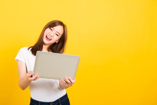 Asian happy portrait beautiful cute young woman teen smile standing wear t-shirt hold laptop computer and excited celebrating success looking to camera, studio shot yellow background with copy space