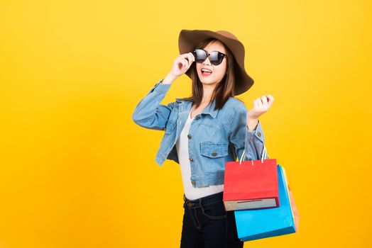 Asian happy portrait beautiful cute young woman teen smiling standing with sunglasses excited holding shopping bags multi color looking camera isolated, studio shot yellow background with copy space
