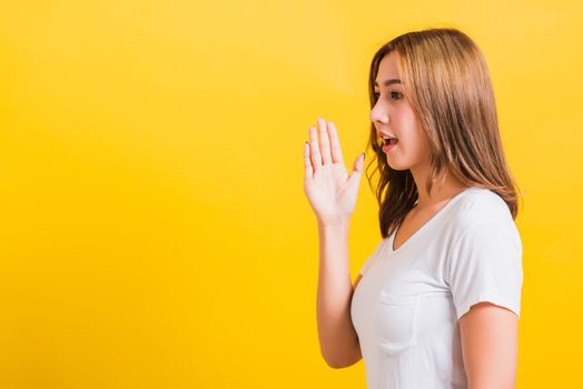 Asian Thai happy portrait beautiful young woman standing wear t-shirt hold a hand on mouth talking whispering secret rumor looking to camera isolated, studio shot on yellow background with copy space