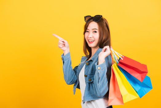 Asian happy portrait beautiful cute young woman teen smiling standing with sunglasses excited holding shopping bags multi color pointing to side isolated, studio shot yellow background with copy space