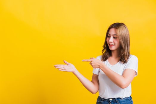 Asian Thai happy portrait beautiful young woman standing to hold something on palm away side and point the finger to it looking to side, studio shot isolated on yellow background with copy space