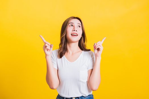 Asian Thai happy portrait beautiful cute young woman standing wear white t-shirt makes gesture two fingers point upwards above looking above, studio shot isolated on yellow background with copy space