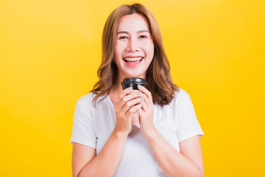 Portrait Asian Thai beautiful happy young woman wear white t-shirt standing smiling holding take away coffee paper cup, studio shot isolated on yellow background, with copy space