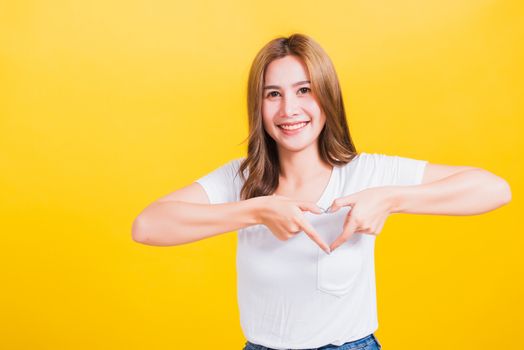 Asian Thai happy portrait beautiful cute young woman smile standing make finger heart figure symbol shape sign with two hands and looking camera, studio shot isolated yellow background with copy space