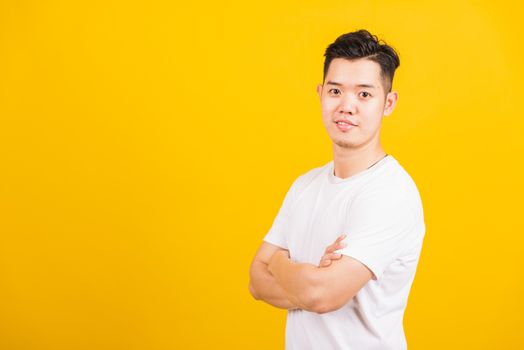 Portrait happy Asian handsome young man smiling standing wearing white t-shirt folded or crossed arm he looking to camera, studio shot isolated yellow background