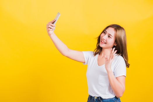 Asian Thai portrait happy beautiful young woman smiling making selfie photo or video call on smartphone she raise hand to say hello to smartphone, studio shot isolated yellow background and copy space